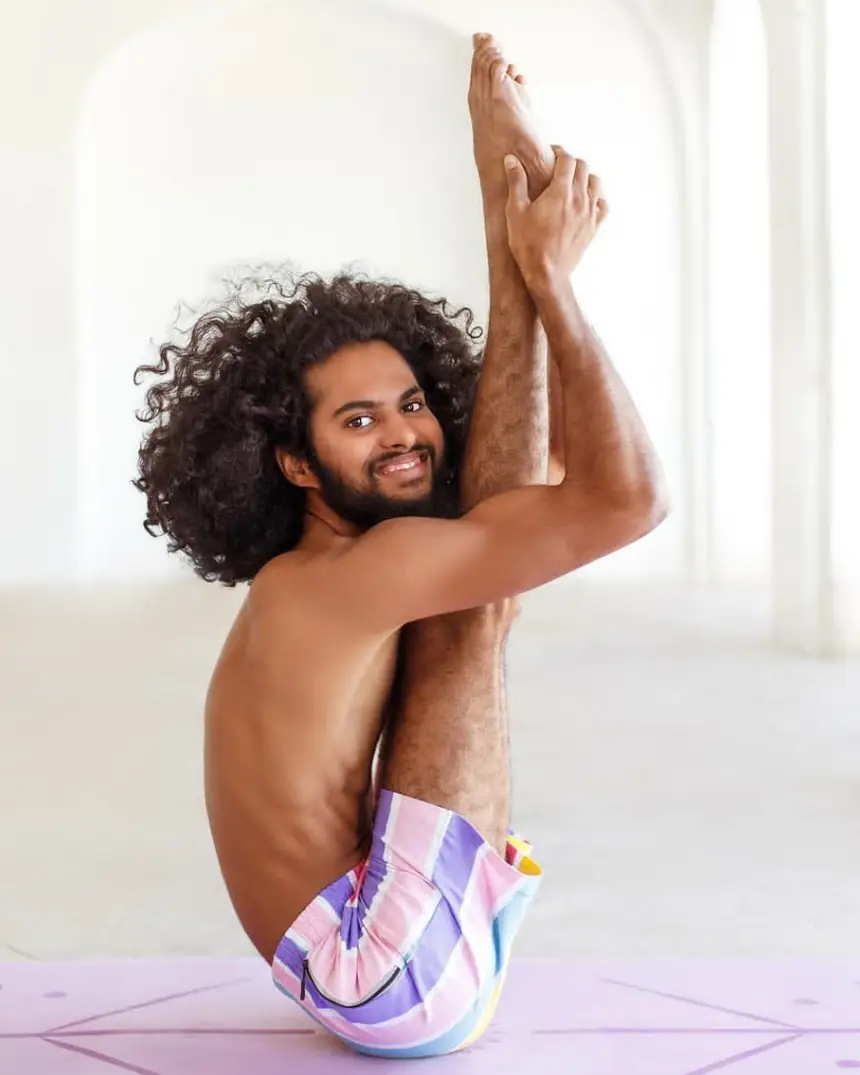 Nithin smiling from a seated compression pose, one leg lifted straight beside his head