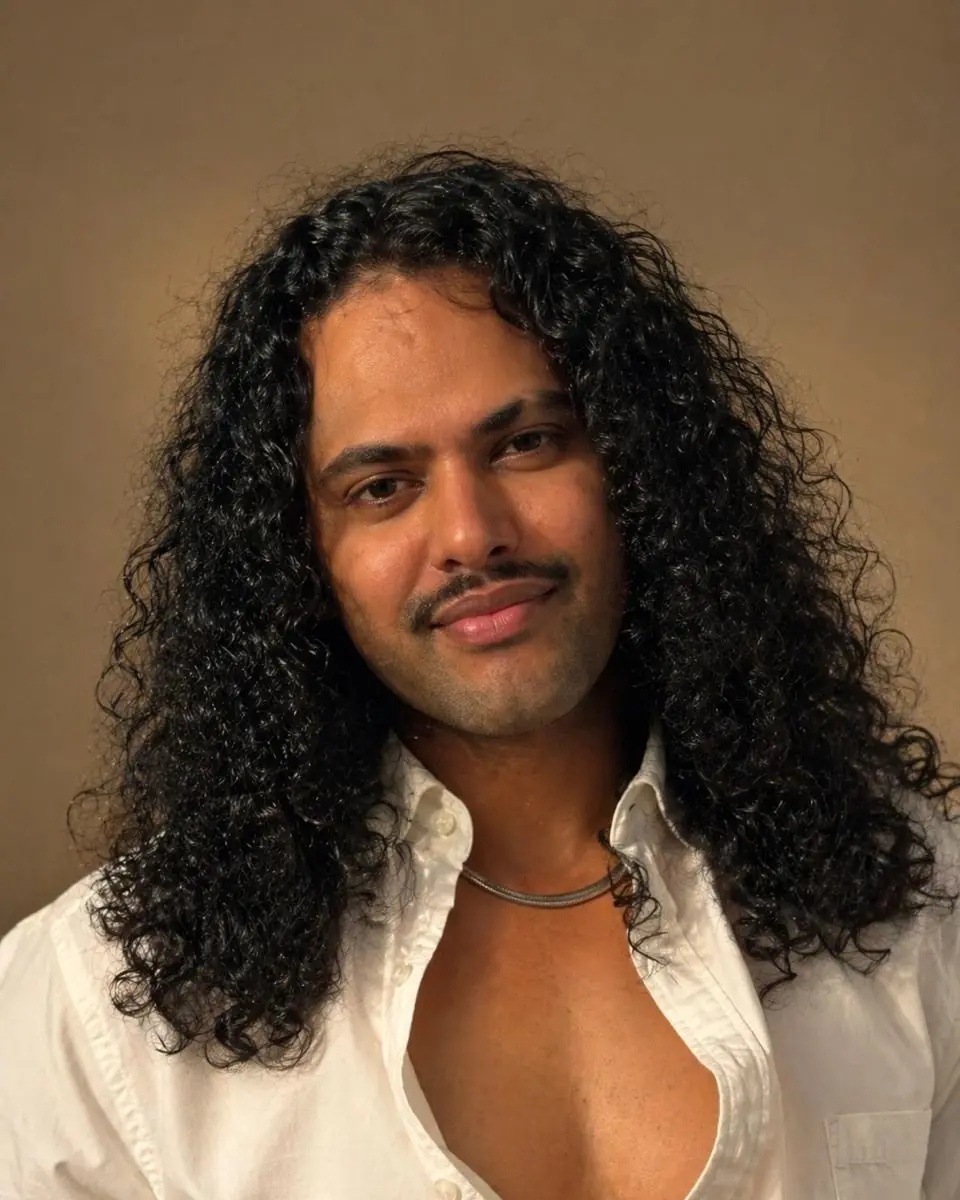 Portrait of Nithin Xavier, long curly hair over an open white shirt, smiling softly against a warm backdrop